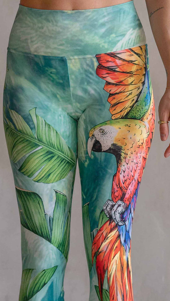 Model wearing WERKSHOP Parrot Athleisure Leggings. The artwork on the leggings feature a tropical parrot down the wearer's left leg and bright green and teal floral artwork on the right leg.