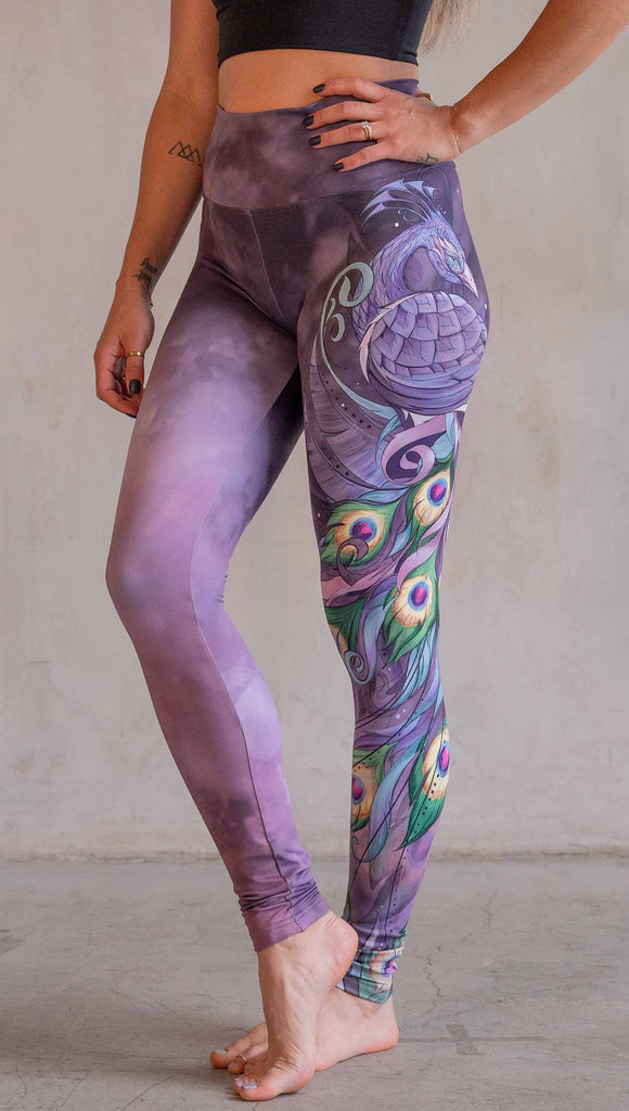 Model wearing WERKSHOP Peacock Athleisure leggings. The leggings have original artwork of a peacock on the wearer's left leg with the body of the bird a the hip and peacock feathers cascading down the leg. The background (and right leg) have a solid purple airbrushed background