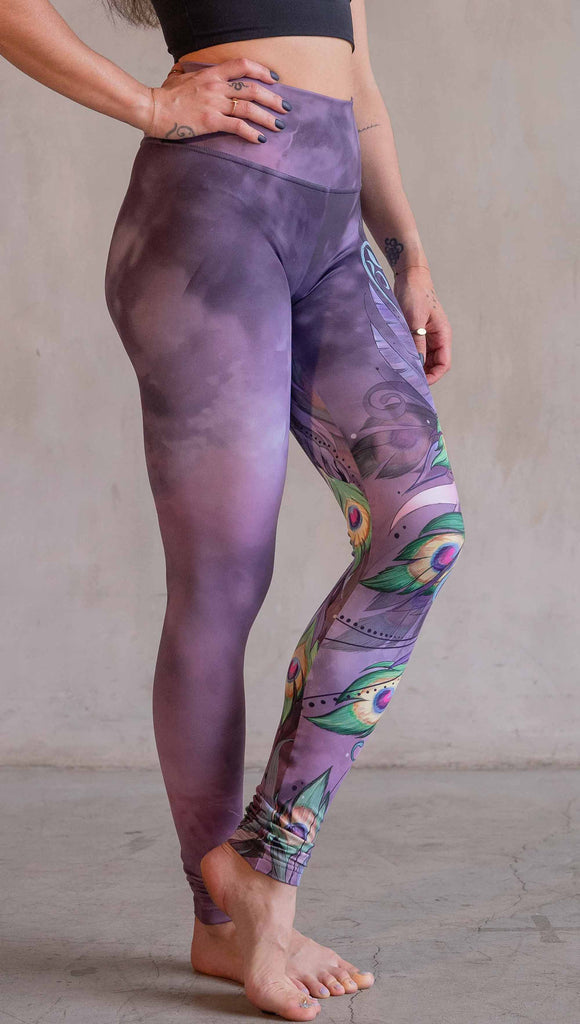 Model wearing WERKSHOP Peacock Athleisure leggings. The leggings have original artwork of a peacock on the wearer's left leg with the body of the bird a the hip and peacock feathers cascading down the leg. The background (and right leg) have a solid purple airbrushed background