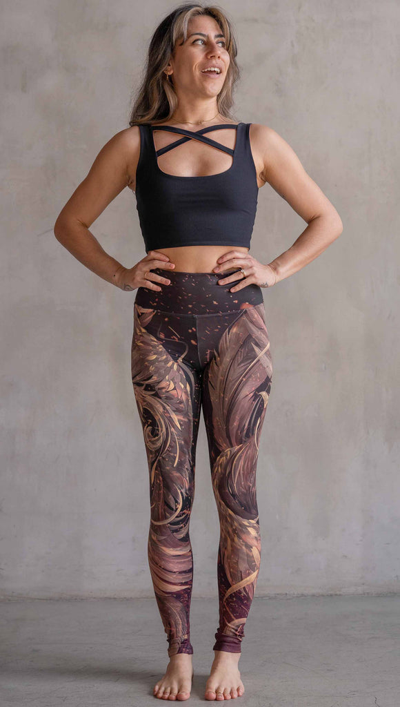 Model wearing WERKSHOP Phoenix Athleisure Leggings. The leggings are printed with a mythical phoenix swirling up each leg with embers flying through the background. The overall color theme is super earthy and warm browns, oranges and yellow tones