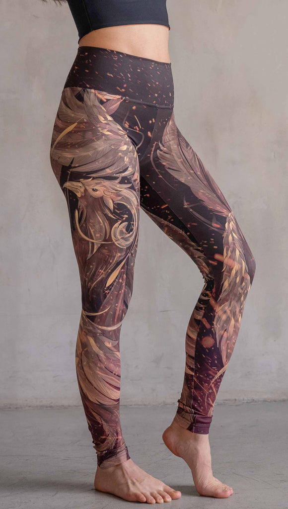 Model wearing WERKSHOP Phoenix Athleisure Leggings. The leggings are printed with a mythical phoenix swirling up each leg with embers flying through the background. The overall color theme is super earthy and warm browns, oranges and yellow tones