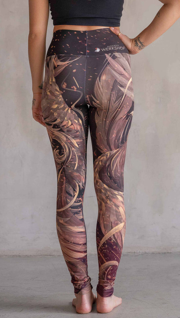 Model wearing WERKSHOP Phoenix Athleisure Leggings. The leggings are printed with a mythical phoenix swirling up each leg with embers flying through the background. The overall color theme is super earthy and warm browns, oranges and yellow tones