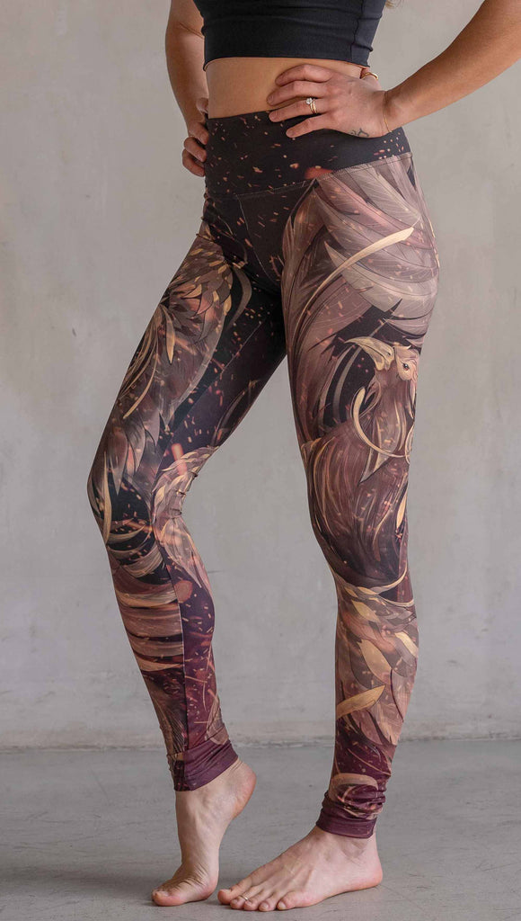 Model wearing WERKSHOP Phoenix Athleisure Leggings. The leggings are printed with a mythical phoenix swirling up each leg with embers flying through the background. The overall color theme is super earthy and warm browns, oranges and yellow tones