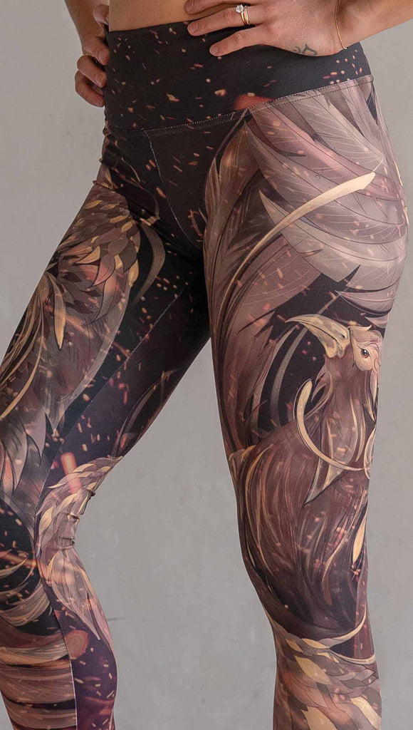 Model wearing WERKSHOP Phoenix Athleisure Leggings. The leggings are printed with a mythical phoenix swirling up each leg with embers flying through the background. The overall color theme is super earthy and warm browns, oranges and yellow tones