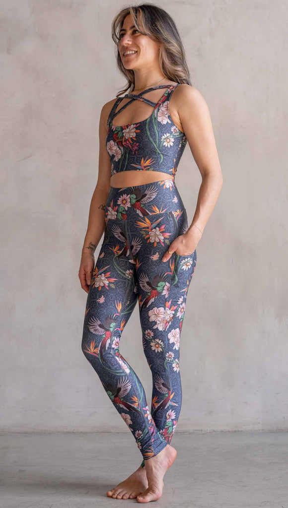 Model wearing WERKSHOP Quetzal Featherlight Leggings. The leggings are printed with with original quetzal artwork with clusters of tropical flowers and birds of paradise over a blue background. The featherlight leggings have pockets.