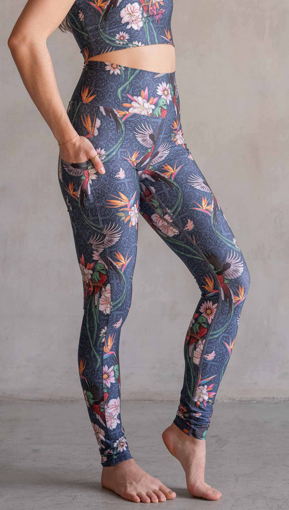 Model wearing WERKSHOP Quetzal Featherlight Leggings. The leggings are printed with with original quetzal artwork with clusters of tropical flowers and birds of paradise over a blue background. The featherlight leggings have pockets.