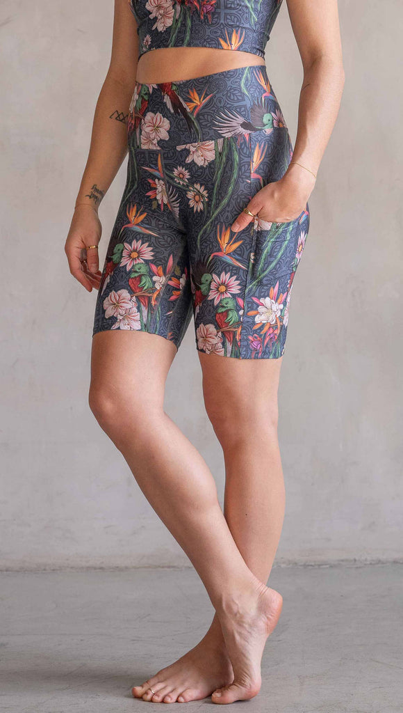 Model wearing WERKSHOP Quetzal Featherlight Bicycle length shorts. The shorts are printed with with original quetzal artwork with clusters of tropical flowers and birds of paradise over a blue background. The featherlight shirt have pockets.