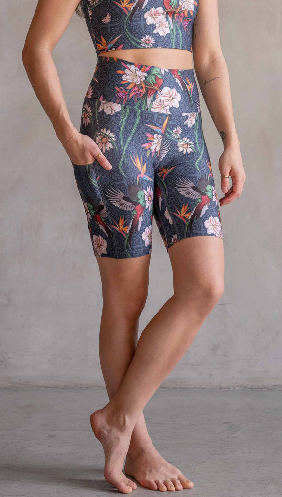 Model wearing WERKSHOP Quetzal Featherlight Bicycle length shorts. The shorts are printed with with original quetzal artwork with clusters of tropical flowers and birds of paradise over a blue background. The featherlight shirt have pockets.