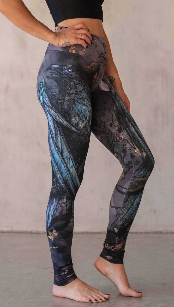 Girl wearing WERKSHOP "Raven" athleisure leggings. The artwork on the leggings feature a raven perched on a branch looking into the moonlight with pops of blue glistening from his feathers. He is framed by autumn leaves over a very spooky night sky.