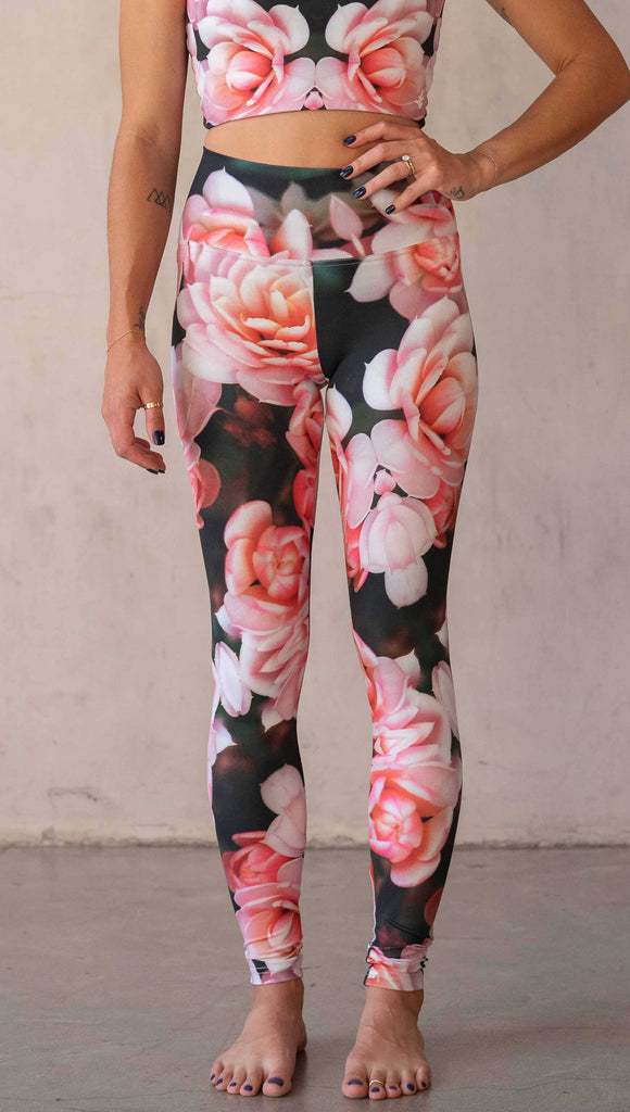 Girl wearing WERKSHOP Rosaline Floral Athleisure Leggings. The leggings feature a fun geometric floral print. It has bright pink flowers with pops of bright peach/orange hues. The flowers are over a super dark blackish greenish background