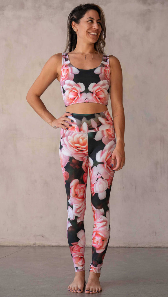 Girl wearing WERKSHOP Rosaline Floral Athleisure Leggings. The leggings feature a fun geometric floral print. It has bright pink flowers with pops of bright peach/orange hues. The flowers are over a super dark blackish greenish background