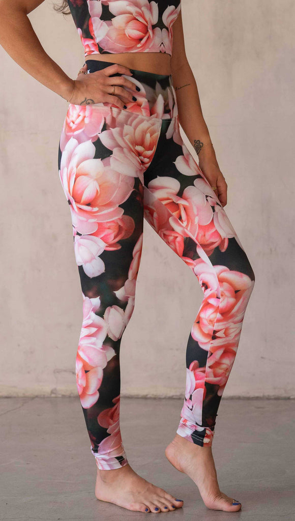 Girl wearing WERKSHOP Rosaline Floral Athleisure Leggings. The leggings feature a fun geometric floral print. It has bright pink flowers with pops of bright peach/orange hues. The flowers are over a super dark blackish greenish background