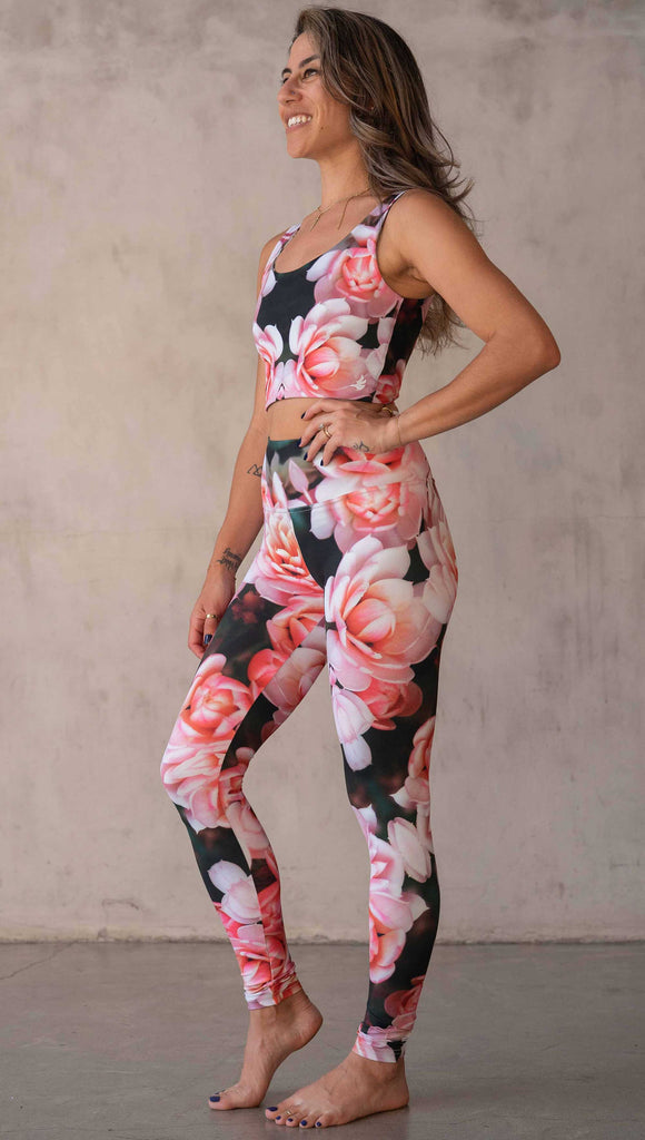 Girl wearing WERKSHOP Rosaline Floral Athleisure Leggings. The leggings feature a fun geometric floral print. It has bright pink flowers with pops of bright peach/orange hues. The flowers are over a super dark blackish greenish background