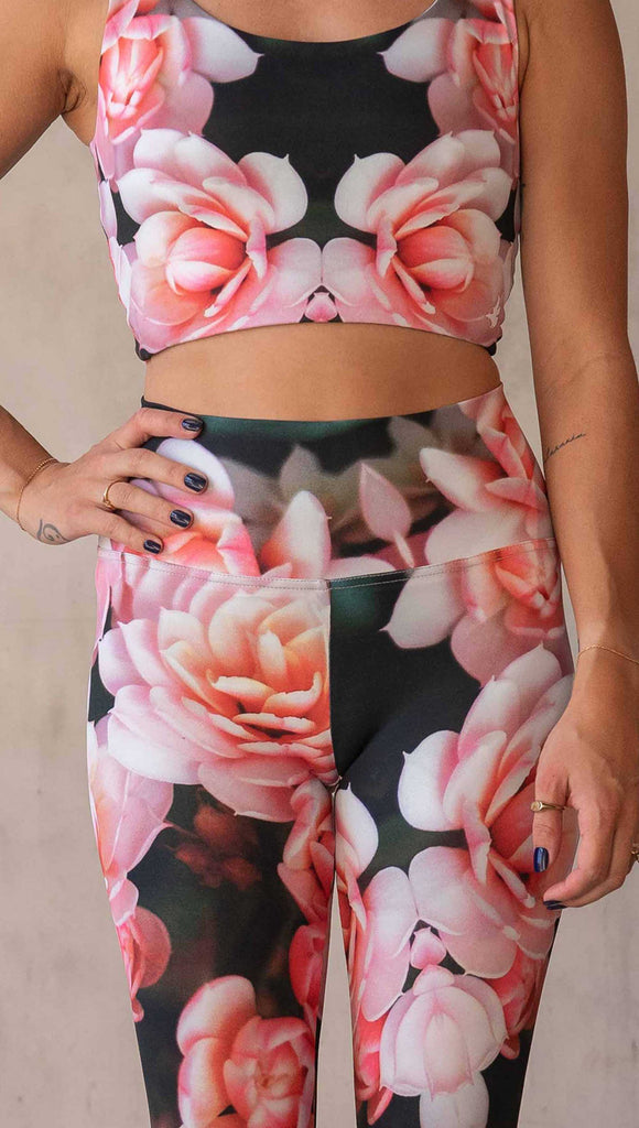 Girl wearing WERKSHOP Rosaline Floral Athleisure Leggings. The leggings feature a fun geometric floral print. It has bright pink flowers with pops of bright peach/orange hues. The flowers are over a super dark blackish greenish background