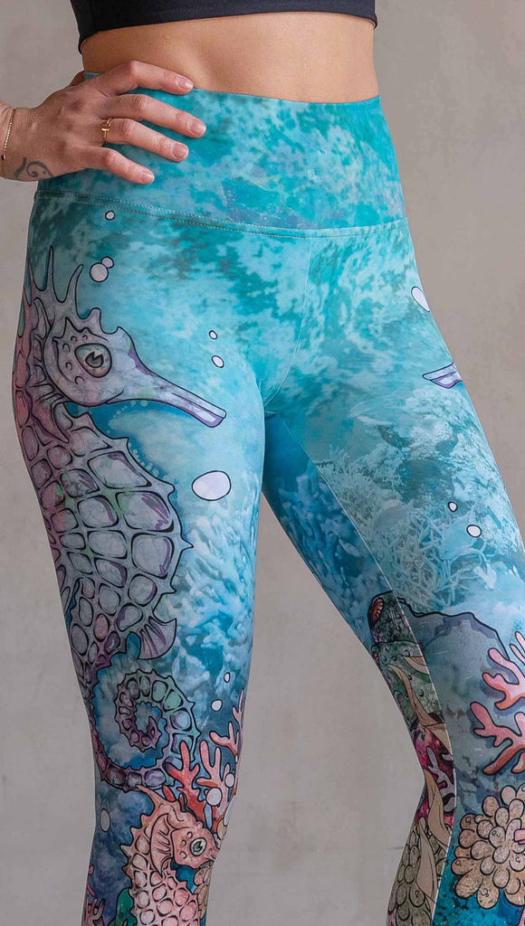 Model wearing WERKSHOP Seahorses Athleisure Leggings. The artwork on the leggings features pastel coloured seahorses and coral reef over an aqua/teal textured background.