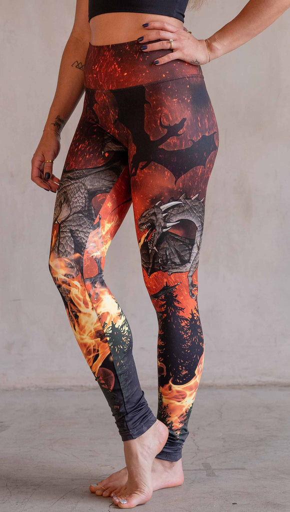 Model wearing WERKSHOP Spitfire Leggings. The artwork on the leggings features a detailed fire breathing dragon blowing flames into a forest with a bright red sky filled with floating embers and silhouettes of more dragons flying in the background.