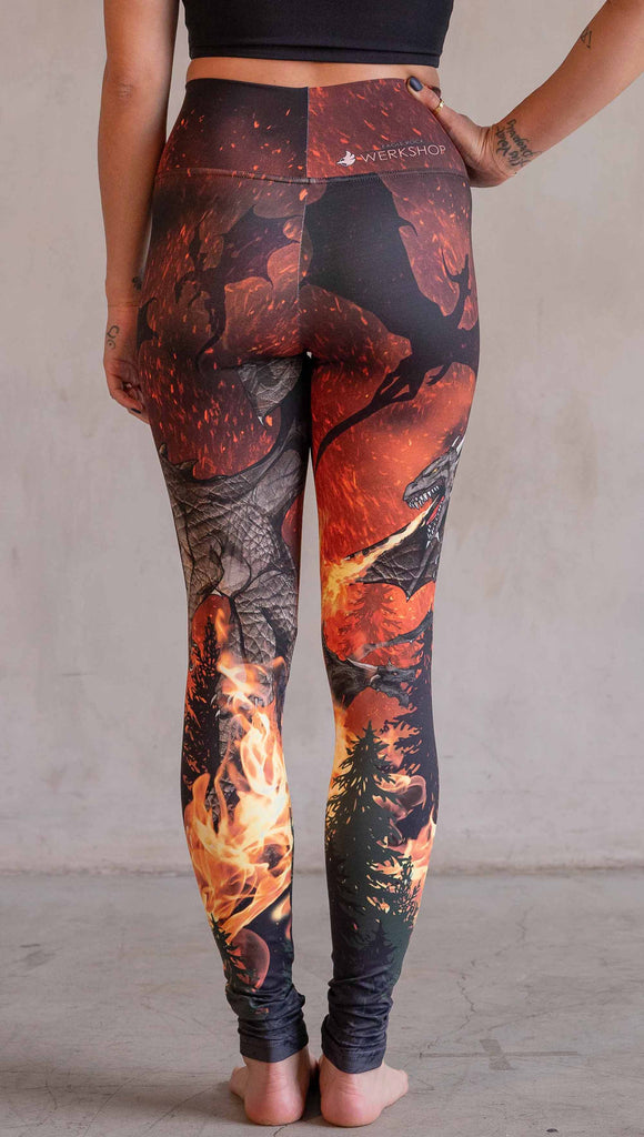 Model wearing WERKSHOP Spitfire Leggings. The artwork on the leggings features a detailed fire breathing dragon blowing flames into a forest with a bright red sky filled with floating embers and silhouettes of more dragons flying in the background.