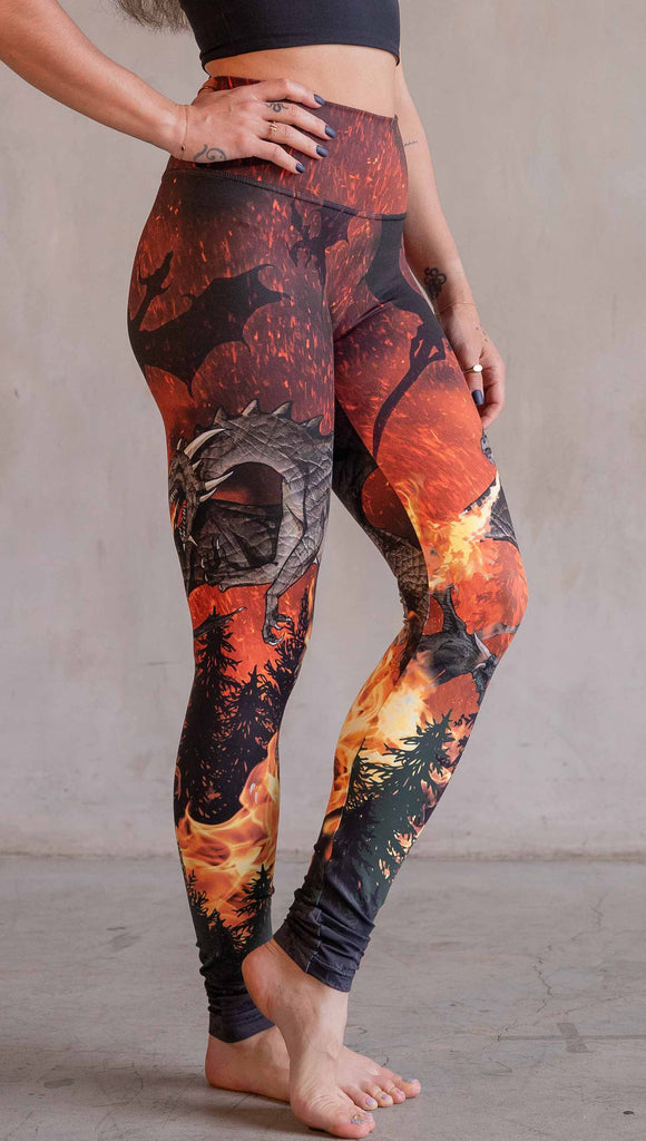 Model wearing WERKSHOP Spitfire Leggings. The artwork on the leggings features a detailed fire breathing dragon blowing flames into a forest with a bright red sky filled with floating embers and silhouettes of more dragons flying in the background.