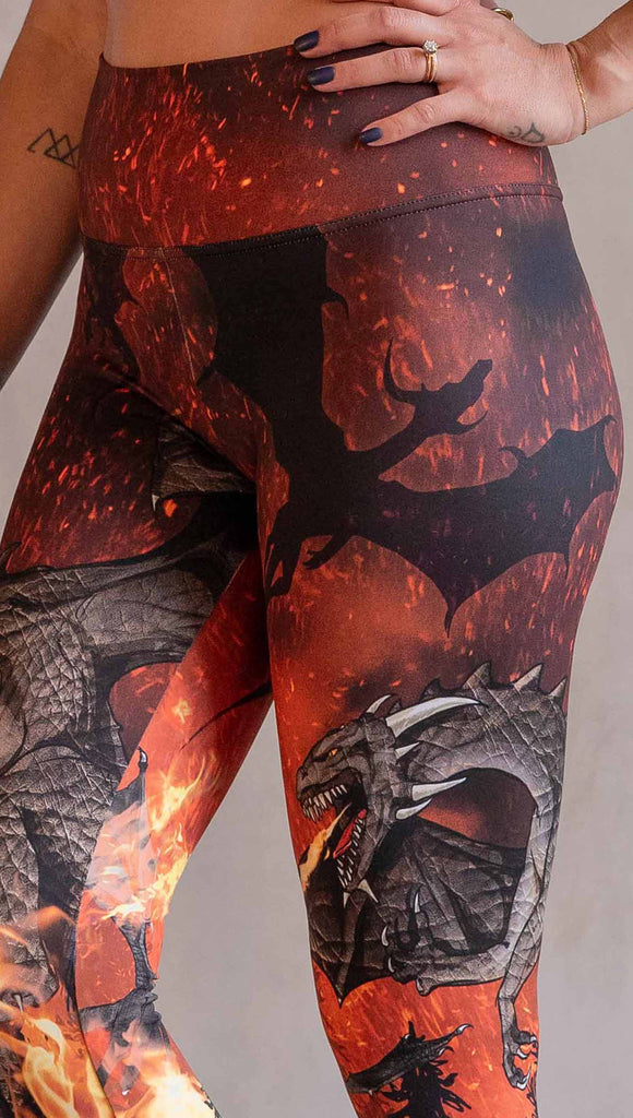 Model wearing WERKSHOP Spitfire Leggings. The artwork on the leggings features a detailed fire breathing dragon blowing flames into a forest with a bright red sky filled with floating embers and silhouettes of more dragons flying in the background.