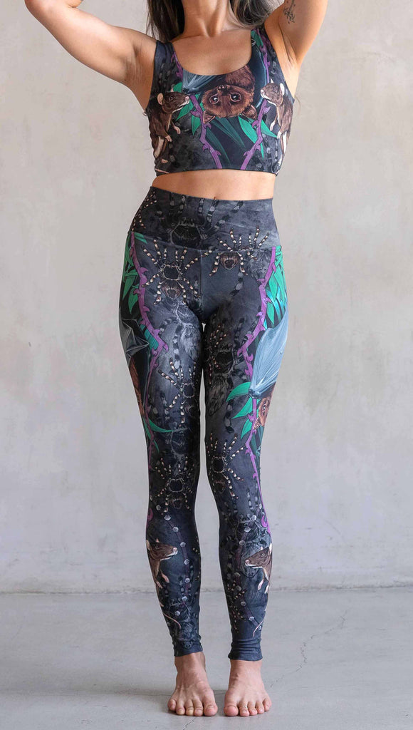 Model wearing WERKSHOP Spooky Season Set. The leggings feature an adorable fruit  bat dangling upside down inside a tropical scene with a purple wreath of thorns. under the bat, there is a rat facing forward. The background is a distressed dark gray brushstroke texture with scattered tarantulas.