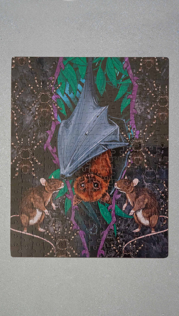 WERKSHOP Spooky Season jigsaw puzzle The artwork features an adorable fruit bat dangling upside down inside a tropical scene with a purple wreath of thorns. Under the bat, there are two rats. The background is a distressed dark gray brushstroke texture. The puzzle comes with a canvas drawcord pouch.