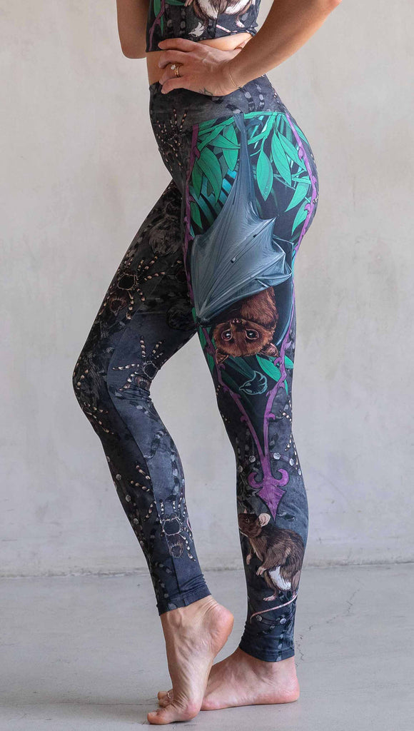 Model wearing WERKSHOP Spooky Season Set. The leggings feature an adorable fruit  bat dangling upside down inside a tropical scene with a purple wreath of thorns. under the bat, there is a rat facing forward. The background is a distressed dark gray brushstroke texture with scattered tarantulas.