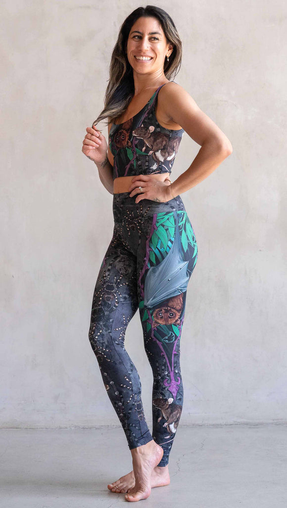 Model wearing WERKSHOP Spooky Season Set. The leggings feature an adorable fruit  bat dangling upside down inside a tropical scene with a purple wreath of thorns. under the bat, there is a rat facing forward. The background is a distressed dark gray brushstroke texture with scattered tarantulas.