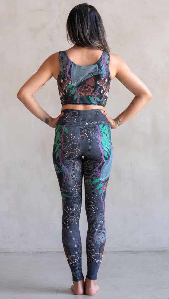 Model wearing WERKSHOP Spooky Season Set. The leggings feature an adorable fruit  bat dangling upside down inside a tropical scene with a purple wreath of thorns. under the bat, there is a rat facing forward. The background is a distressed dark gray brushstroke texture with scattered tarantulas.