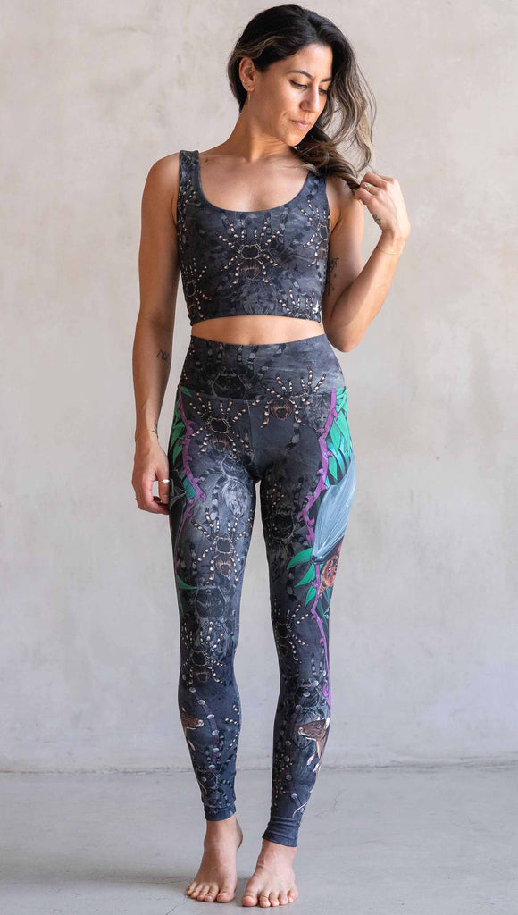 Model wearing WERKSHOP Spooky Season Set. The leggings feature an adorable fruit  bat dangling upside down inside a tropical scene with a purple wreath of thorns. under the bat, there is a rat facing forward. The background is a distressed dark gray brushstroke texture with scattered tarantulas.