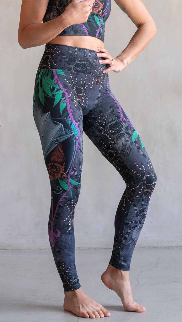 Model wearing WERKSHOP Spooky Season Set. The leggings feature an adorable fruit bat dangling upside down inside a tropical scene with a purple wreath of thorns. The background is a distressed dark gray brushstroke texture. (This version of the artwork has no rats)