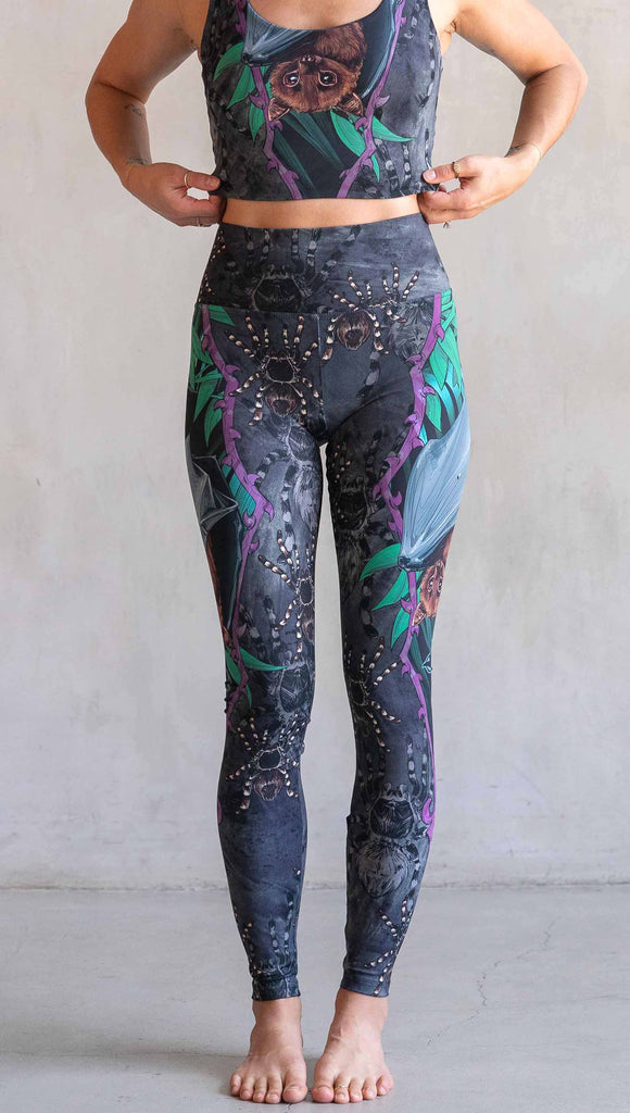 Model wearing WERKSHOP Spooky Season Set. The leggings feature an adorable fruit bat dangling upside down inside a tropical scene with a purple wreath of thorns. The background is a distressed dark gray brushstroke texture. (This version of the artwork has no rats)