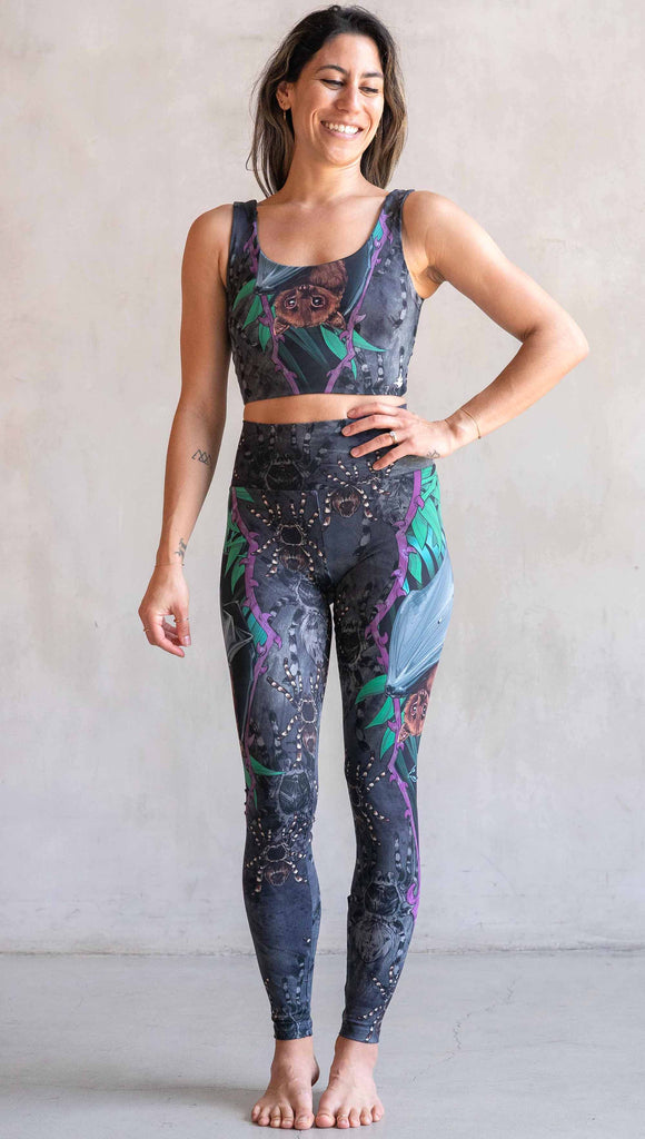 Model wearing WERKSHOP Spooky Season Set. The leggings feature an adorable fruit bat dangling upside down inside a tropical scene with a purple wreath of thorns. The background is a distressed dark gray brushstroke texture. (This version of the artwork has no rats)