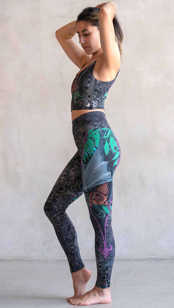 Model wearing WERKSHOP Spooky Season Set. The leggings feature an adorable fruit bat dangling upside down inside a tropical scene with a purple wreath of thorns. The background is a distressed dark gray brushstroke texture. (This version of the artwork has no rats)