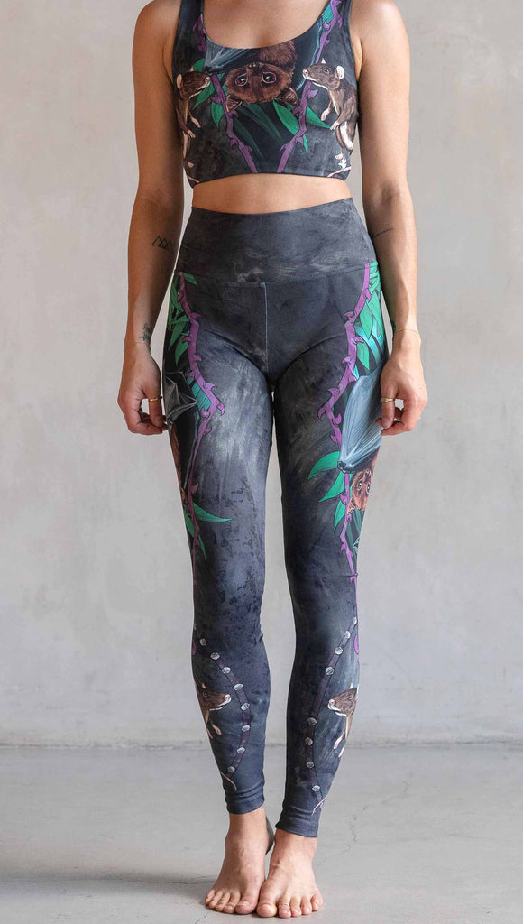 Model wearing WERKSHOP Spooky Season Set. The leggings feature an adorable fruit bat dangling upside down inside a tropical scene with a purple wreath of thorns. under the bat, there is a rat facing forward. The background is a distressed dark gray brushstroke texture. (This version of the artwork has no spiders)