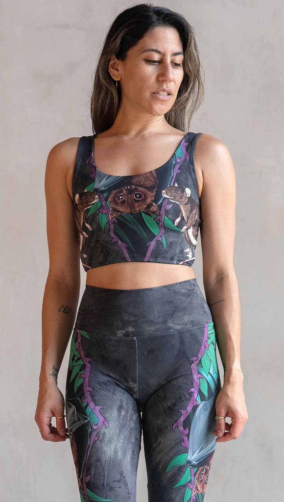 Model wearing WERKSHOP Spooky Season Set. The leggings feature an adorable fruit bat dangling upside down inside a tropical scene with a purple wreath of thorns. under the bat, there is a rat facing forward. The background is a distressed dark gray brushstroke texture. (This version of the artwork has no spiders)