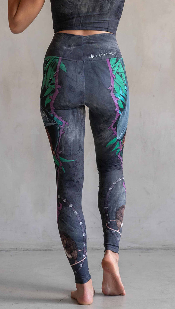 Model wearing WERKSHOP Spooky Season Set. The leggings feature an adorable fruit bat dangling upside down inside a tropical scene with a purple wreath of thorns. under the bat, there is a rat facing forward. The background is a distressed dark gray brushstroke texture. (This version of the artwork has no spiders)