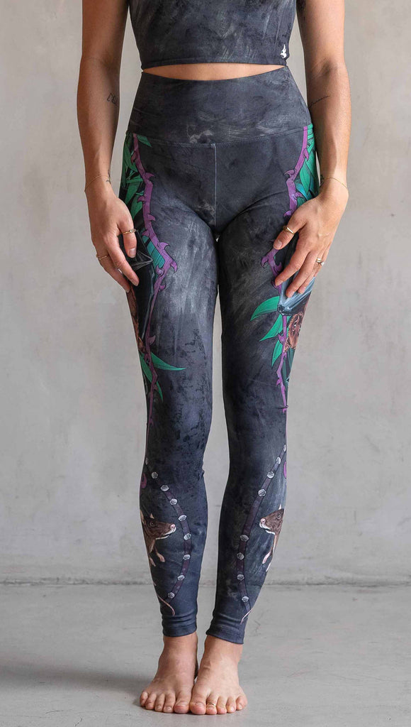 Model wearing WERKSHOP Spooky Season Set. The leggings feature an adorable fruit bat dangling upside down inside a tropical scene with a purple wreath of thorns. under the bat, there is a rat facing forward. The background is a distressed dark gray brushstroke texture. (This version of the artwork has no spiders)