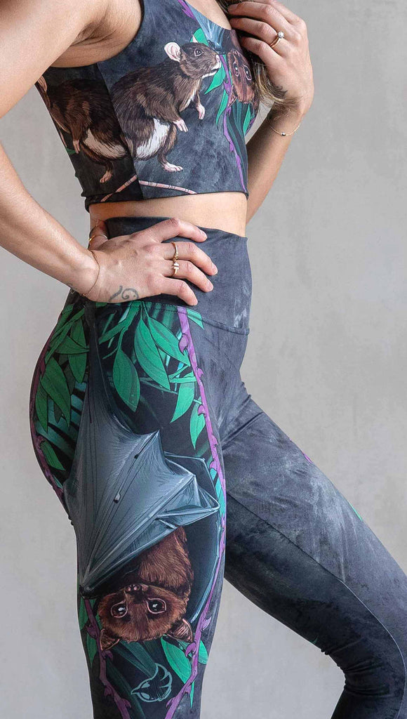 Model wearing WERKSHOP Spooky Season Set. The leggings feature an adorable fruit bat dangling upside down inside a tropical scene with a purple wreath of thorns. under the bat, there is a rat facing forward. The background is a distressed dark gray brushstroke texture. (This version of the artwork has no spiders)