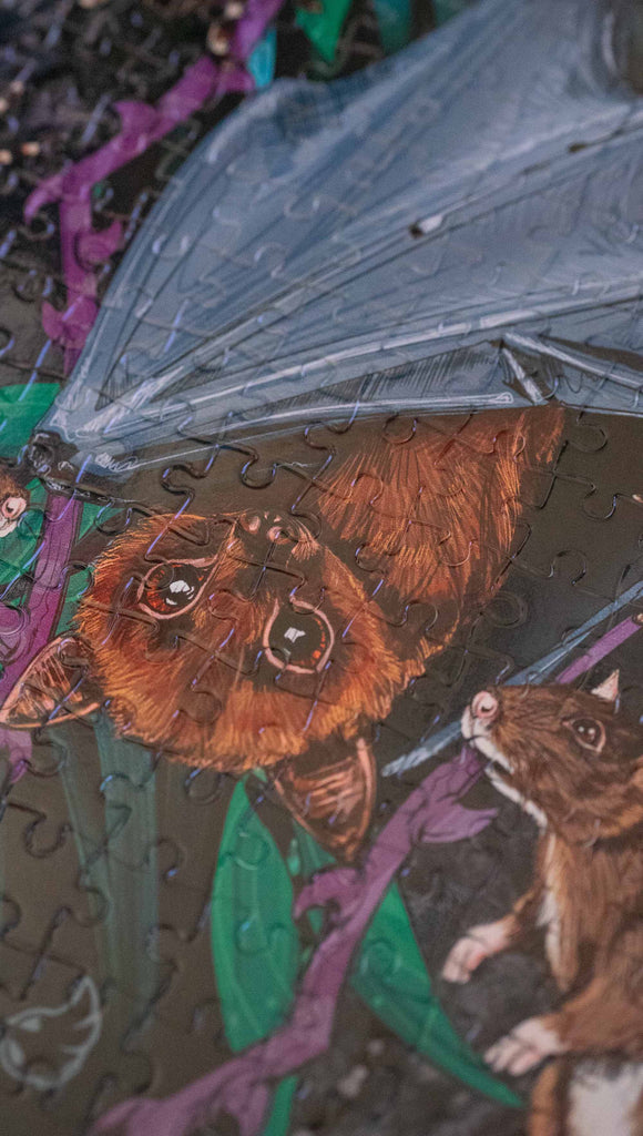 WERKSHOP Spooky Season jigsaw puzzle The artwork features an adorable fruit bat dangling upside down inside a tropical scene with a purple wreath of thorns. Under the bat, there are two rats. The background is a distressed dark gray brushstroke texture. The puzzle comes with a canvas drawcord pouch.