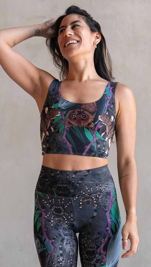 Model wearing WERKSHOP Spooky Season reversible top. One side features an adorable fruit bat dangling upside down inside a tropical scene with a purple wreath of thorns. under the bat, there is a rat facing forward. The background is a distressed dark gray brushstroke texture with scattered tarantulas. The other side is just "ghost" tarantulas.