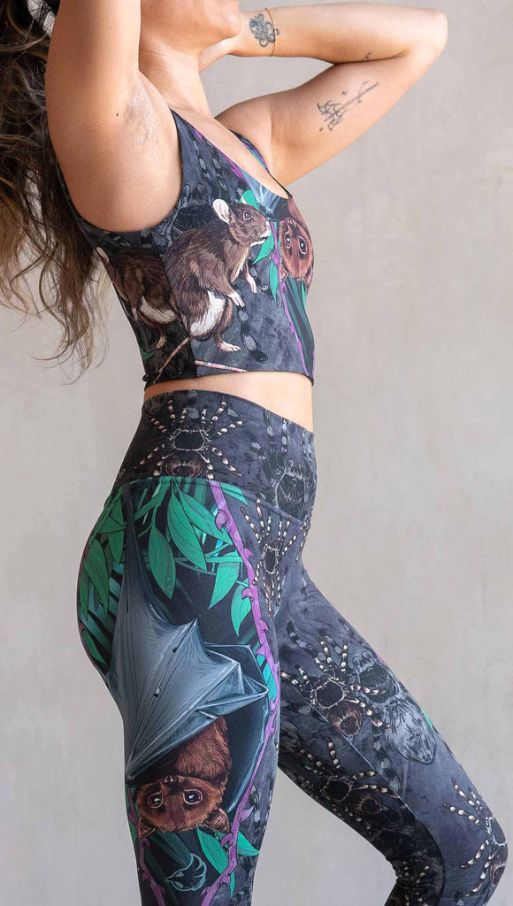 Zoomed in photo of model wearing WERKSHOP Spooky Season Set. The leggings feature an adorable fruit  bat dangling upside down inside a tropical scene with a purple wreath of thorns. under the bat, there is a rat facing forward. The background is a distressed dark gray brushstroke texture with scattered tarantulas.