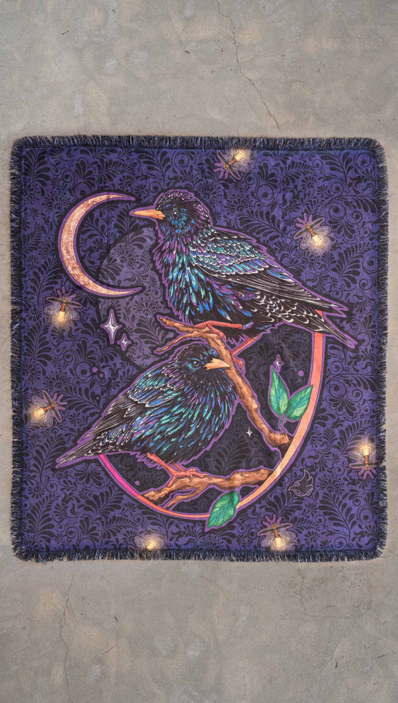 WERKSHOP Starlings tapestry draped on a couch. The tapestry is printed with original artwork by Chriztina Marie fearuring Featuring European Starlings perched on a branch near a crescent moon and fireflies. The colors are warm purples with pops of pink, gold and green.