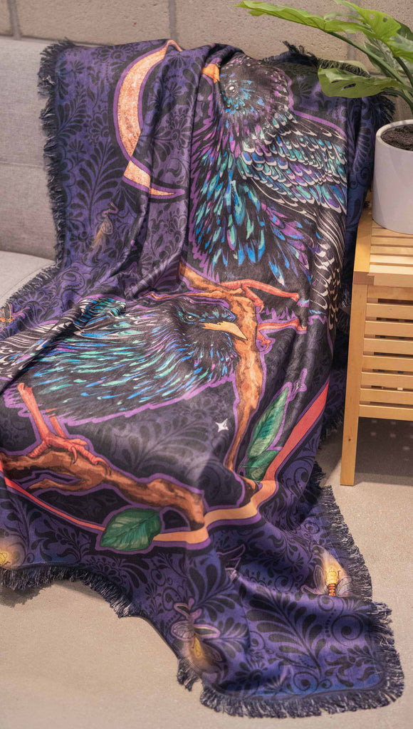 WERKSHOP Starlings tapestry draped on a couch. The tapestry is printed with original artwork by Chriztina Marie fearuring Featuring European Starlings perched on a branch near a crescent moon and fireflies. The colors are warm purples with pops of pink, gold and green.