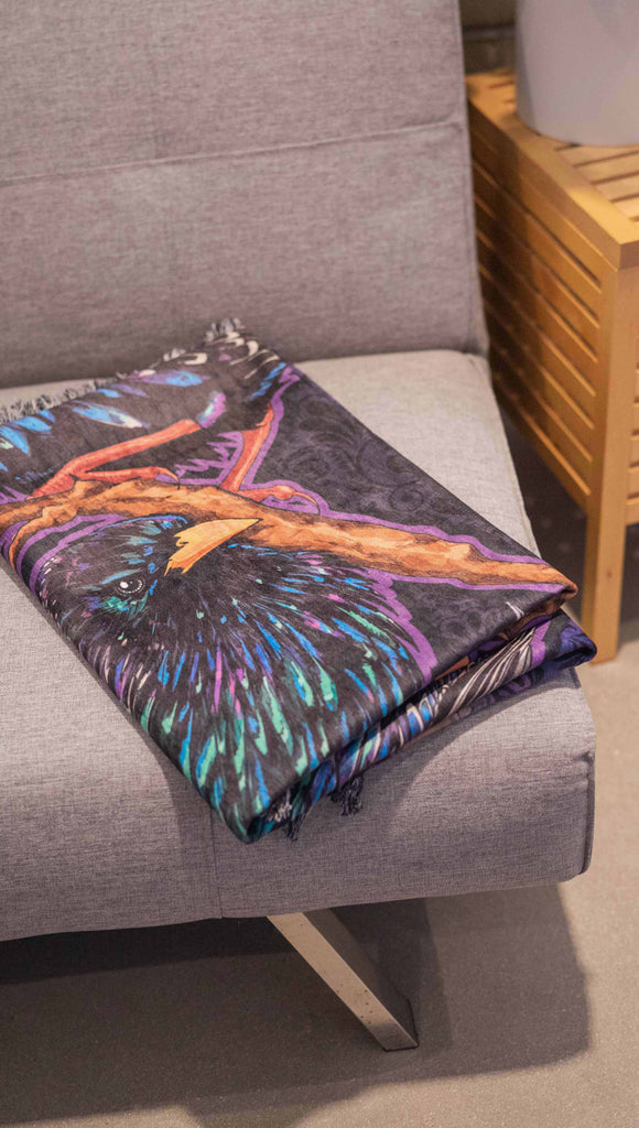 WERKSHOP Starlings tapestry draped on a couch. The tapestry is printed with original artwork by Chriztina Marie fearuring Featuring European Starlings perched on a branch near a crescent moon and fireflies. The colors are warm purples with pops of pink, gold and green.