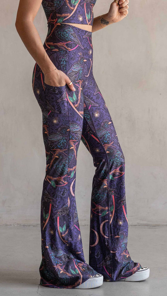 Side view of model wearing WERKSHOP Starlings EnviSoft bell bottom pants with pockets. The fabric is printed with original artwork by our Female Founder, Chriztina Marie. Featuring European Starlings perched on a branch near a crescent moon and fireflies. The colors are warm purples with pops of pink, gold and green.