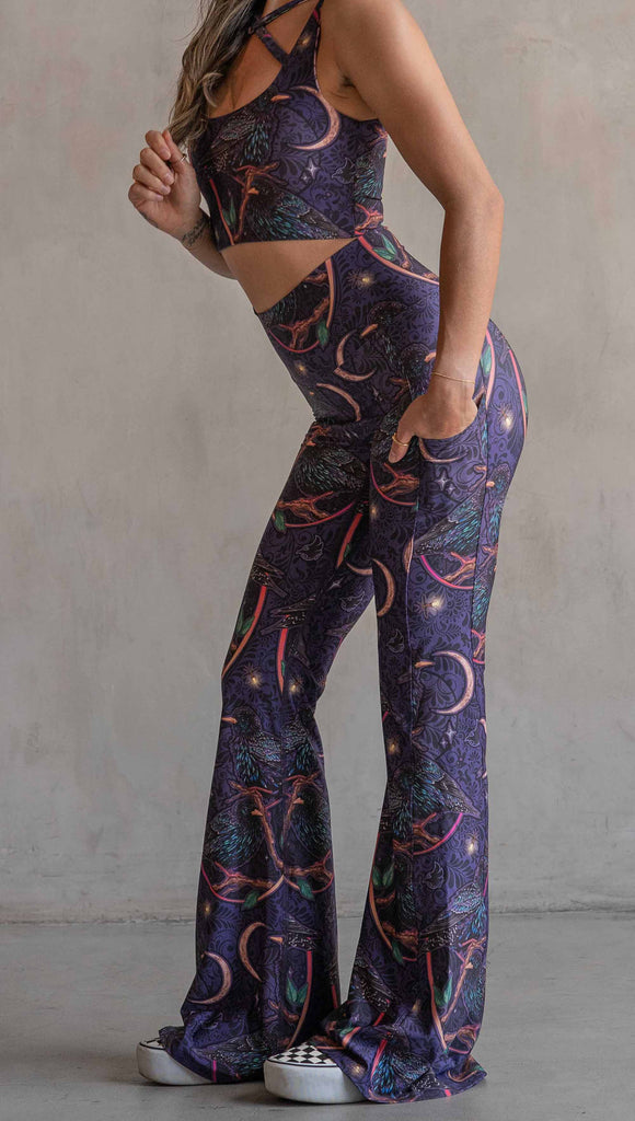 Side view of model wearing WERKSHOP Starlings EnviSoft bell bottom pants with pockets. The fabric is printed with original artwork by our Female Founder, Chriztina Marie. Featuring European Starlings perched on a branch near a crescent moon and fireflies. The colors are warm purples with pops of pink, gold and green.