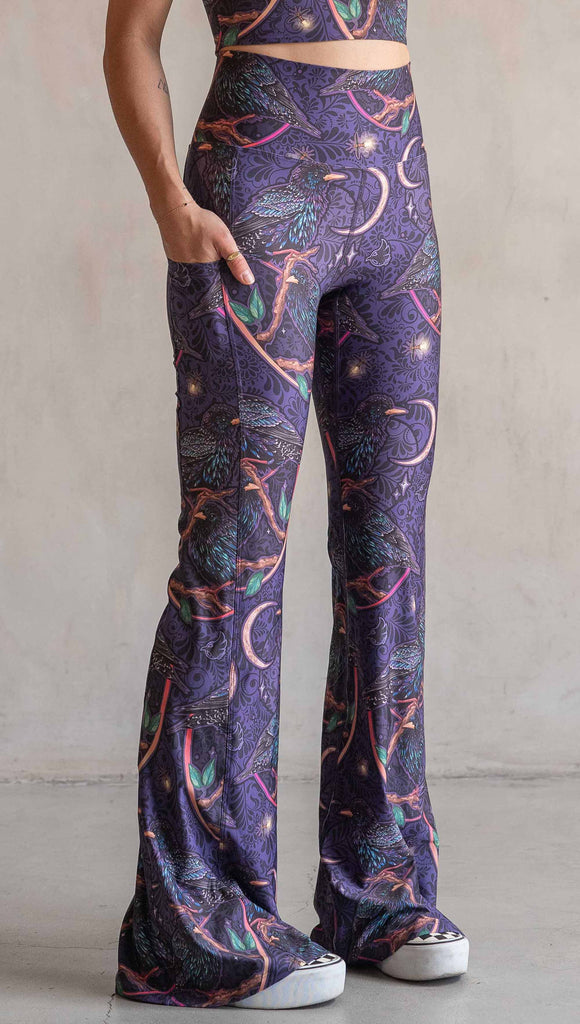 Side view of model wearing WERKSHOP Starlings EnviSoft bell bottom pants with pockets. The fabric is printed with original artwork by our Female Founder, Chriztina Marie. Featuring European Starlings perched on a branch near a crescent moon and fireflies. The colors are warm purples with pops of pink, gold and green.