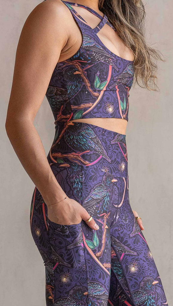 Zoomed in view of model wearing WERKSHOP Starlings EnviSoft bell bottom pants with pockets. The fabric is printed with original artwork by our Female Founder, Chriztina Marie. Featuring European Starlings perched on a branch near a crescent moon and fireflies. The colors are warm purples with pops of pink, gold and green.