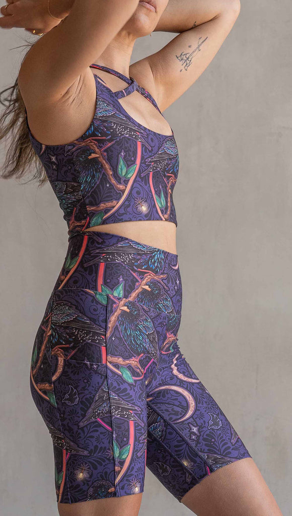 Side view of model wearing WERKSHOP Starlings EnviSoft bicycle length shorts with pockets. The fabric is printed with original artwork by our Female Founder, Chriztina Marie. Featuring European Starlings perched on a branch near a crescent moon and fireflies. The colors are warm purples with pops of pink, gold and green.