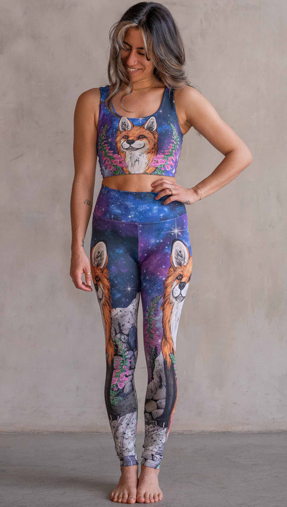 Girl wearing WERKSHOP X Save A Fox Athleisure Leggings. The artwork on the leggings features a coy red fox looking upward while perched on a rock and framed with foxglove flowers. The background for the artwork features a galactic sky with swirls of purple and blue and sparkling stars.